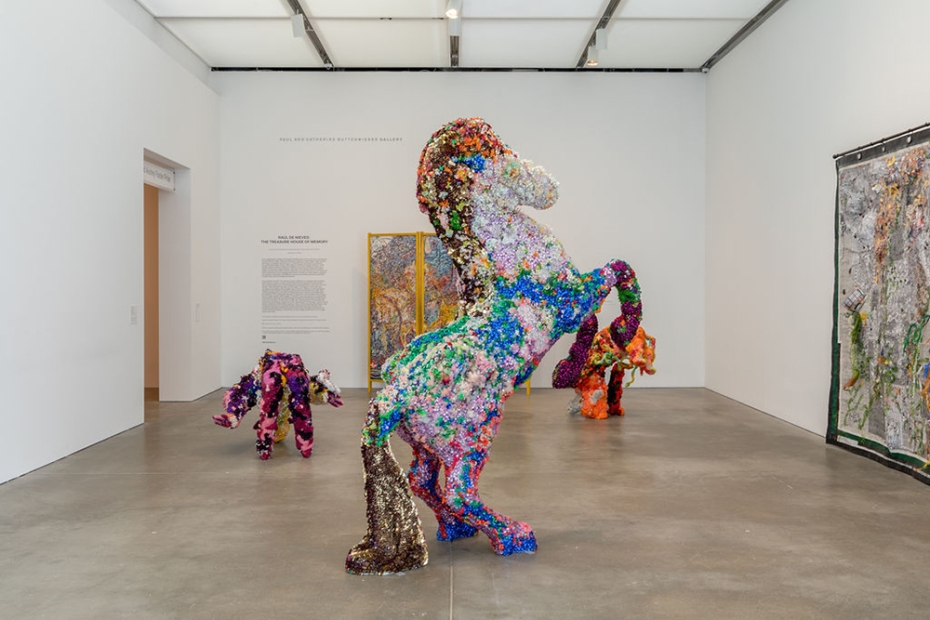 A multi-colored, bejeweled horse sculpture is in the middle of the room. Two smaller, similarly textured sculptures are in view, and a collaged tapestry hangs to the right while a multi-colored folding screen peeks out behind the horse sculpture.