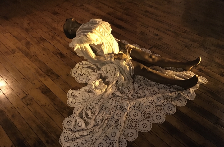 A woman with dark skin wearing a long white lace gown performs on the floor, sitting with her head arched back