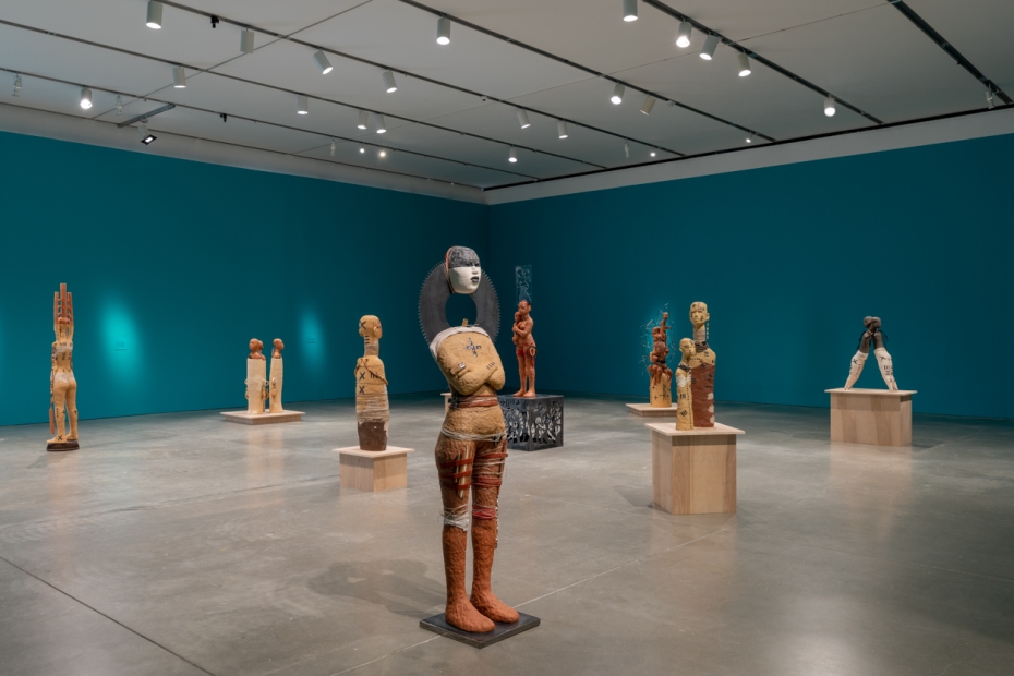 Clay figurative sculptures arranged in an open gallery, some standing on the floor and some on wooden plinths