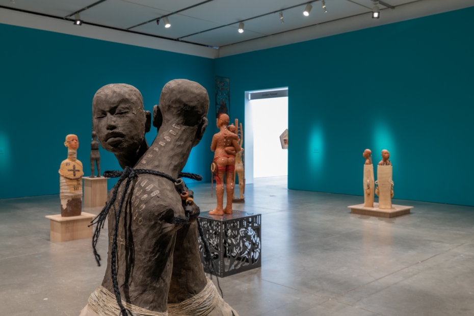 Two interlocked grey figures in front of other clay figures in view in blue gallery