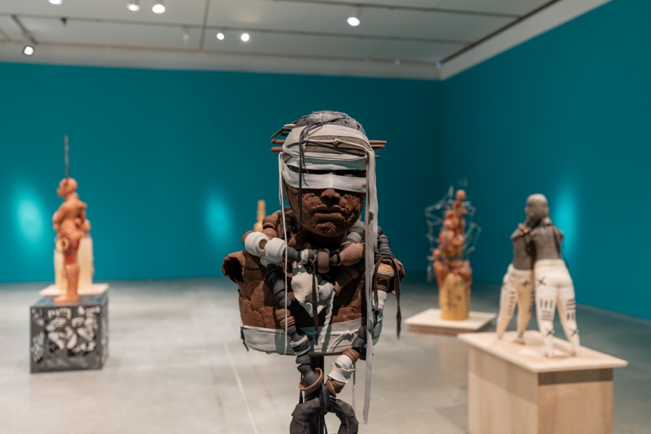 Brown blindfolded clay bust in front of other clay sculptures in blue gallery