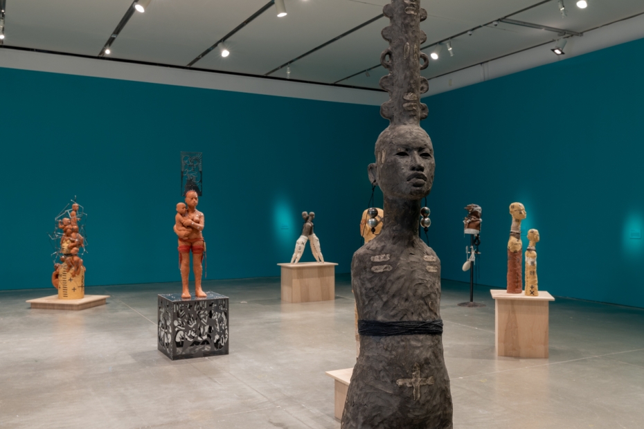 Grey armless figure in front of other clay sculptures in blue gallery