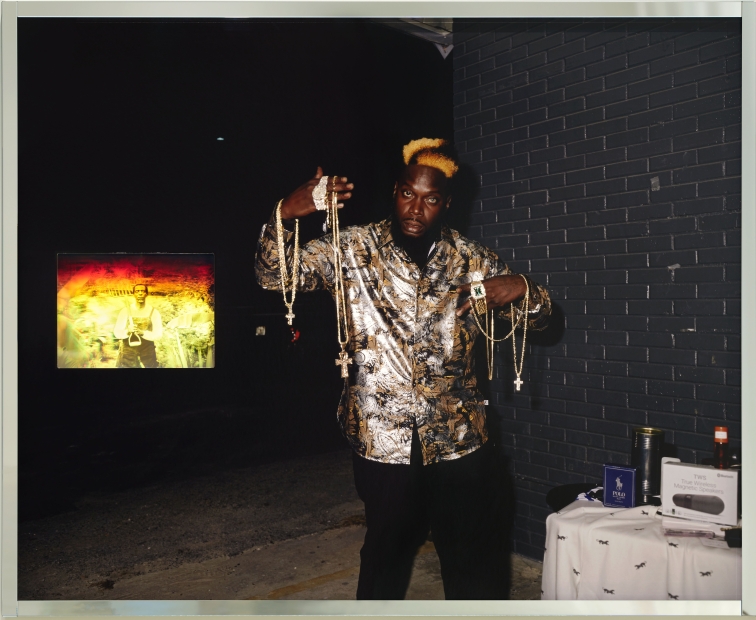 A Black man in a gold and black outfit poses with golden necklaces he is selling, draped from his arms. A hologram of an older Black man in overalls in a farm setting is embedded into the work.