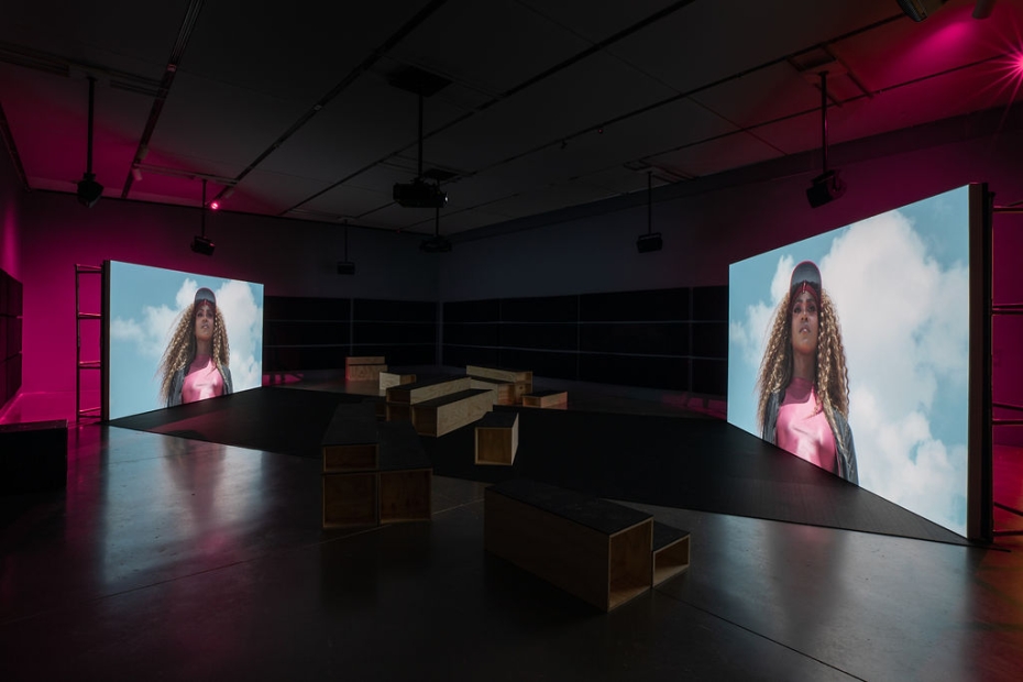 Two large screens displaying a woman with long hair are arranged opposite another in a dark room lit on either side with bright pink light