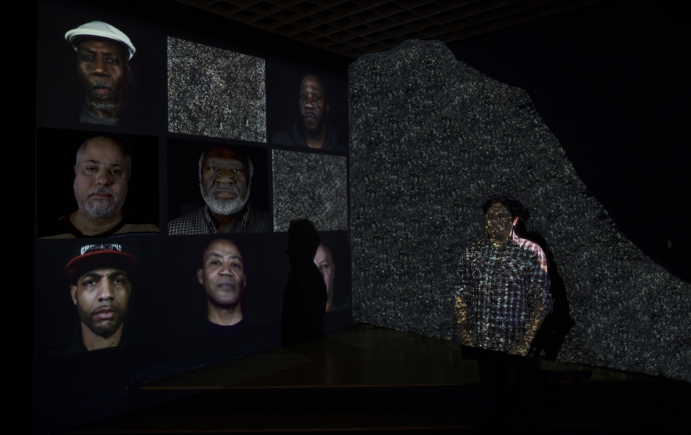 A grid of nine video screens on the left side of the image shows 7 close up portraits of men. Two screens show static. On the right side of the image, a man stands against a wall with his hands in his pockets. He is superimposed with a projection of static.