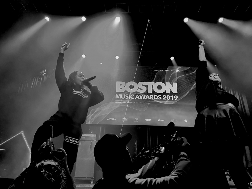 A black and white photo of DJ WhySham and Brandie Blaze performing in front of a banner reading 2019 Boston Music Award.