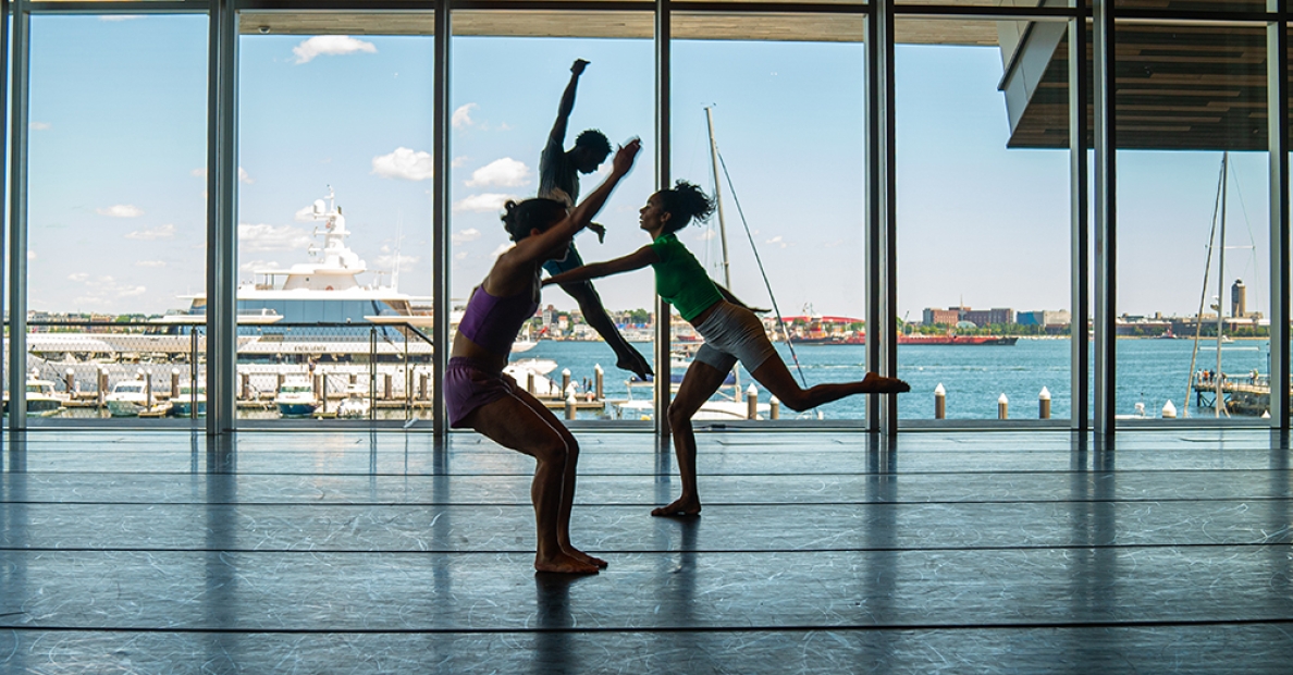 Three dancers backlit against large wall of windows facing the sea
