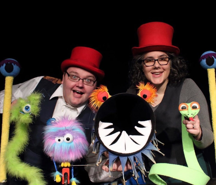 Two puppeteers in red top hats pose with monster puppets