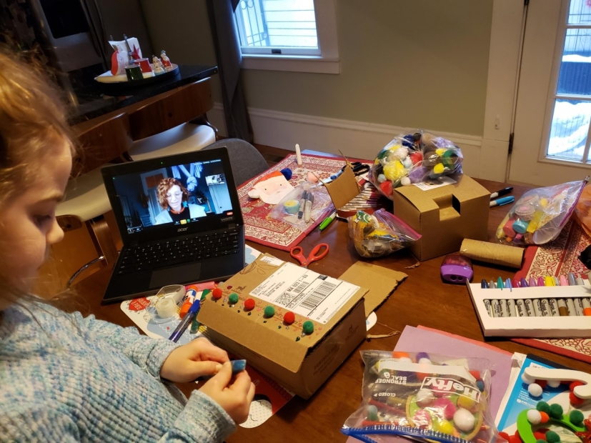 A young child creating an art piece on a dining table with their laptop open and various art supplies.
