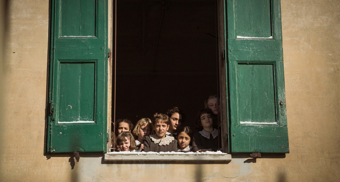 A group of children look out from an open window with green shutters