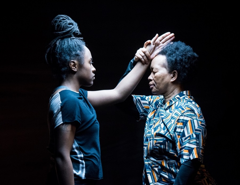 Two Black women face each other before a dark backdrop. The younger stretches an open hand to the head of the older, who grasps her wrist with her eyes closed.