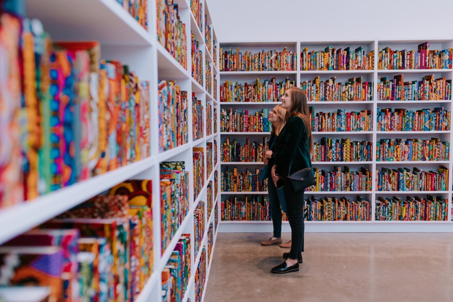 Two women look at walls of books covered in colorful dutch wax fabric, with names embossed in gold on the spines.