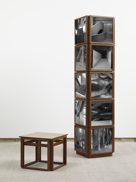 A small wooden stool and a wooden tower made of a stack of cubes with black-and-white photographs of interior staircases. 