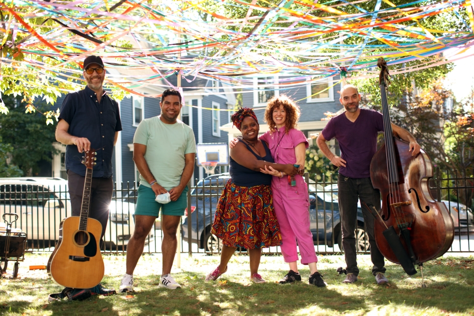 A group of adults pose with a guitar and a string bass outdoors under colorful streamers