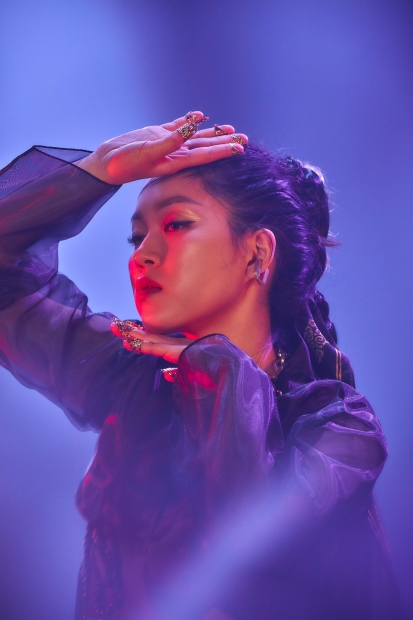 A performer with braided dark hair poses while lit with purple light