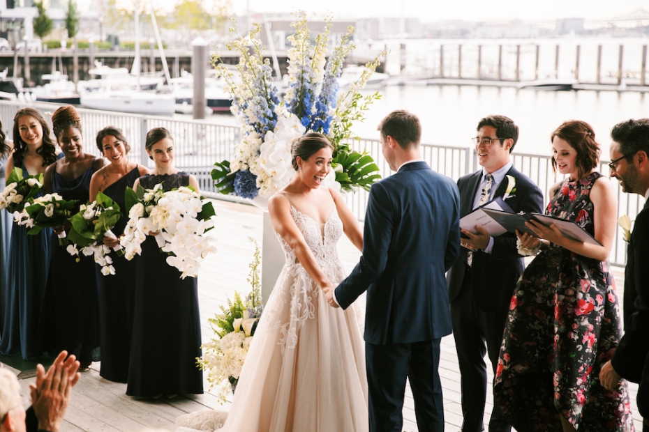 A bride smiles happily at her groom along Boston Harbor as her officiant and wedding party look on.