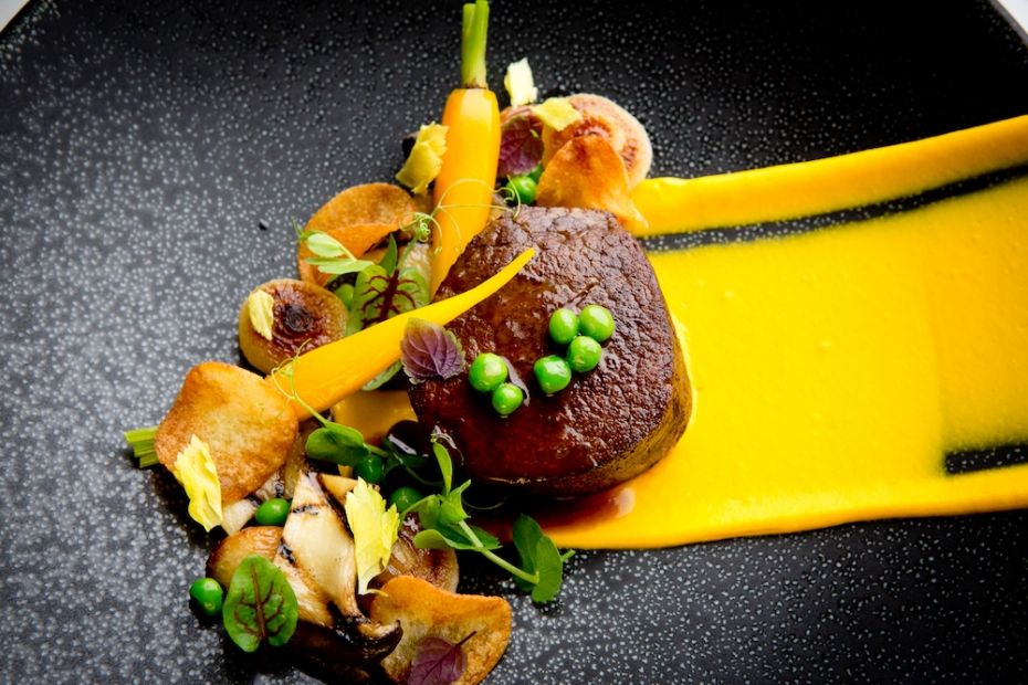 A dish with meat, carrots, peas, and a swipe of orange sauce on a black plate