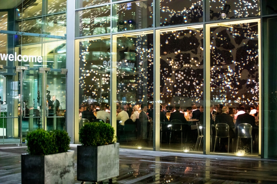 Guests enjoy dinner behind glass windows reflecting hundreds of small lights and bearing the word "welcome".