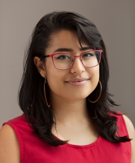 A headshot of Katie Perez wearing a red shirt and red glasses in front of a gray wall.