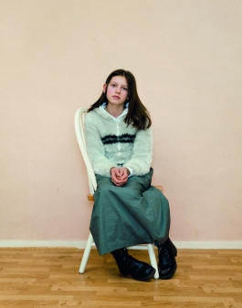 A color photograph of a light-skinned girl with long brown hair wearing a pale blouse and long green skirt sitting sideways on a chair to face the viewer. 