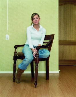 A color photograph of a light-skinned young woman wearing a white blouse and blue jeans, sitting sideways on a chair to face the viewer. 