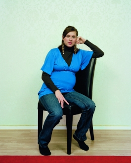 A color photograph of a pregnant light-skinned woman wearing a bright blue blouse and sitting with one arm propped on the back of a black chair, positioned at an angle and gazing at the viewer.