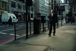 A color photograph of a light-skinned man in a suit walking along an urban street. 