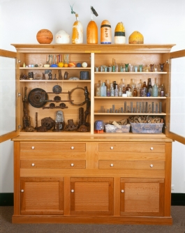 A sculpture of a large, wooden cabinet  with open glass doors displaying a variety of found objects including bottles and buoys. 