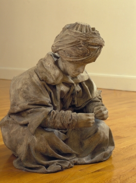 A bronze sculpture of a man in robes and a turban, looking at his hands, seated on the wooden floor of a gallery.