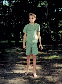 A color photograph of a light-skinned young boy in grayish-green standing in a wooded park. 