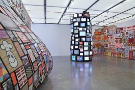 A monumental installation consisting of a towering sculpture made of television screens, a wall-sized mural, and a convex sculpture made of colorful framed graphics coming off the wall.