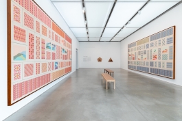 Large, framed embroidered work consisting of patterned rectangles hang opposing each other in a gallery with benches between them