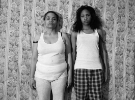 A black-and-white photograph shows the artist, a Black woman, standing shoulder to shoulder with her mother before floral-patterned wallpaper. They are wearing white tank tops and casual bottoms and gaze directly at the viewer.