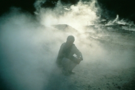 A color photograph shows the shadowy image of a figure crouching in a cloud of illuminated smoke in a hilly landscape.