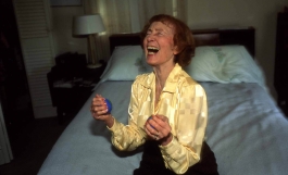 A color photograph of an older light-skinned woman wearing a yellow blouse and black pants and laughing widely while seated on the edge of a bed.