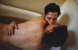 A color photograph of two light-skinned men embracing in a full bathtub as they kiss.