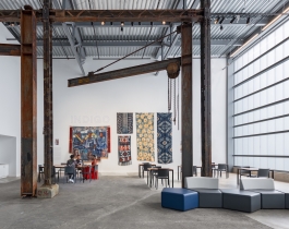 A room with white walls, iron beams and gantries, a selection of colorful fabric wall hangings under the title "INDIGO," and three people sitting at a small table.