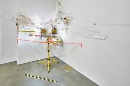 An elaborate mixed media installation occupying the corner of a white room and consisting of peeling white material, various yellow and orange materials, and a yellow metal tripod.