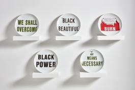 An installation of five ceramic plates displayed on five shelves and printed with the sayings"We Shall Overcome,", "Black is Beautiful,", "Burn Baby Burn,", "Black Power,", and "By Any Means Necessary."