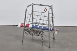 A sculpture made of an orthopedic walker and various salvaged metal works with a rolled up American flag wrapped in plastic laying across it.