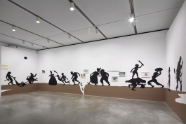 A photograph of an installation comprising black, life-size silhouettes on a brown ground acting out a series of scenes across three walls of a gallery.