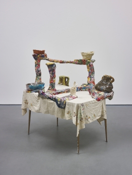 A sculpture consisting of a colorful ceramic and papier mache structure with multiple arms holding ceramic vessels and displayed on a table with an embroidered white tablecloth.