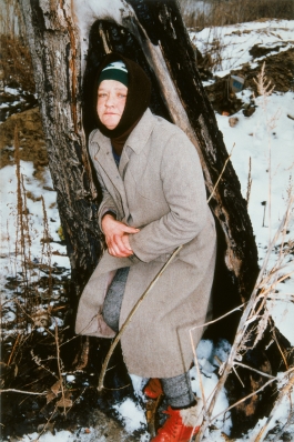 A color photograph shows a pale-skinned figure in a hat, hood, overcoat, and red boots hunched against a tree in a snowy forest, looking directly at the viewer.
