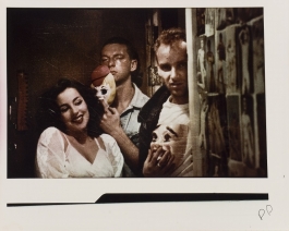 A grainy color photograph shows three light-skinned people in a hallway next to a wall of pinned-up images of nudes: a woman made up like a movie starlet and coyly posed, and two men in open collared shirts, one with a T-shirt underneath, holding theatrical masks and looking directly at the viewer.