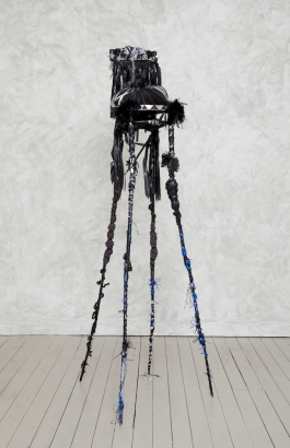 A sculpture comprised of a black wooden chair with elongated, spider-like legs that towers in height and is decorated with black feathers, black and silver tinsel, and hair.