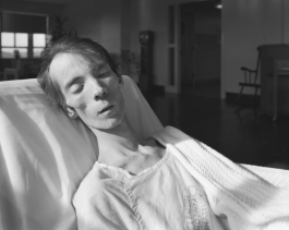 A black-and-white photograph of a pale emaciated man asleep in a hospital bed with bright sunlight shining on his face.