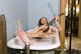 A video still shows the artist, a white man, in a bathtub, wearing headphones, and plays the guitar, and apparently nude.