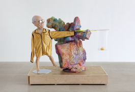 A mixed-media sculpture of a figure built from a Dick Cheney face mask, sunglasses, and a gold jacket along with an amorphous, colorful sculpture on a wooden plinth 