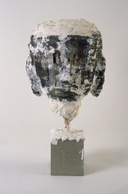 A sculpture of an abstracted, figurative head comprised of mixed materials and suspended upright on a display plinth.