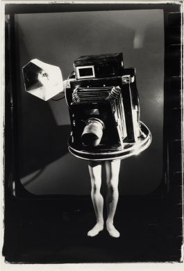 A black-and-white photograph of a person wearing a full-body costume of an antique press camera on a photo set with only their pale exposed legs visible.